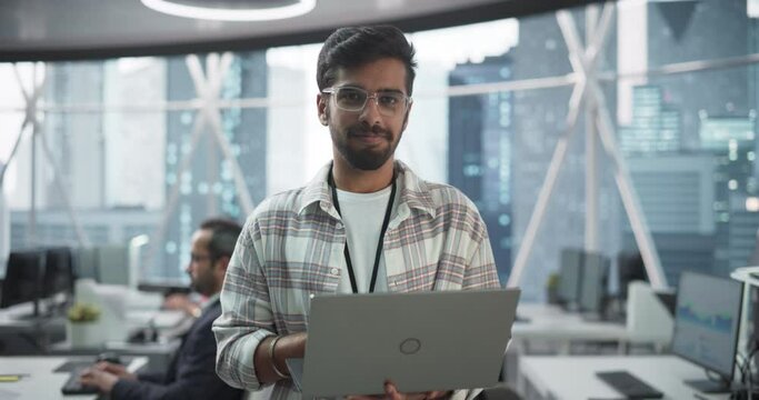 Handsome Indian Man Wearing Glasses, Charmingly Smiling and Looking at Camera. Young Intelligent Male Engineer Holding a Laptop Computer. Finance Manager Working in Modern Corporate Office