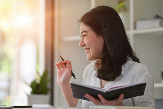 Close up image of Young Asian businesswoman in smart casual outfit smiling and holding a notebook.