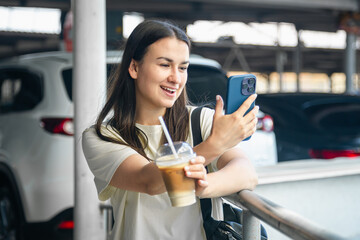 A young woman with a cup of coffee in the parking lot takes a selfie.