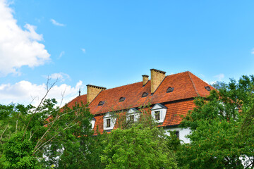 Fototapeta na wymiar Rural house with white walls and red tiled roof in green trees on a sunny summer day with blue sky. Poland, Kurnik, Poznan, June 2022.