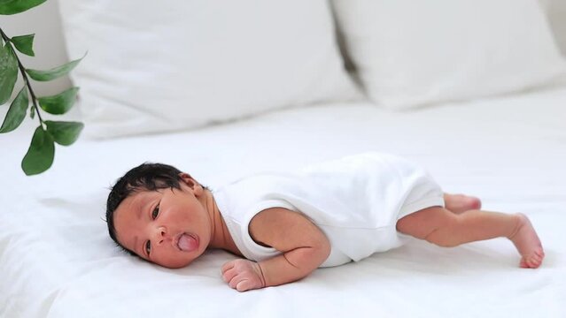 portrait of a newborn black African-American baby funny showing tongue, a small dark-skinned baby close-up lying on the bed on his tummy in the bedroom waking up or falling asleep in a white bodysuit