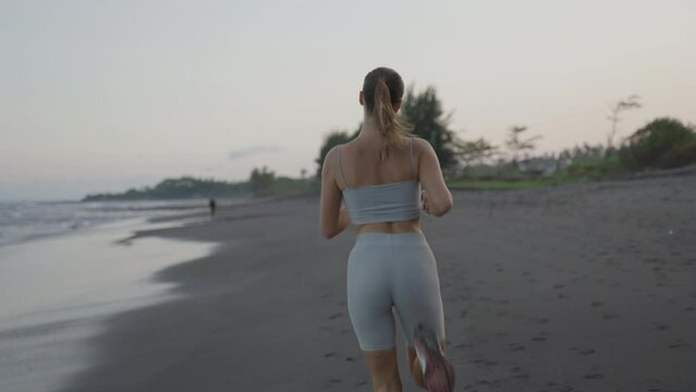 Back view sportswoman running at seashore with dramatic sky and bright sunshine slow motion. Female jogger during outdoor activity on beach. Medium shot 