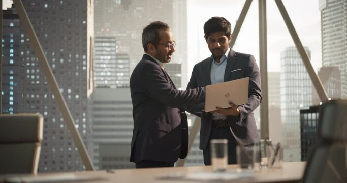 Two Young South Asian Businessmen Standing Next to a Window in a Modern Office with a City Skyline View. Indian Managers Using Laptop Computer, Discussing Business Investments and Market Opportunities