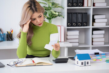 oung Asian woman sitting and calculating expenses and mortgage with calculator and house on table Worried about payments and non-repayable loans, business, and property