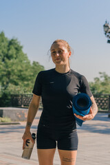 Fototapeta premium portrait of a young smiling sportswoman girl holding a kettlebell and a bottle of water in her hands before starting a workout outdoors in the park