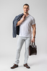 A man in casual clothes holds a leather briefcase and a jacket in his hands.