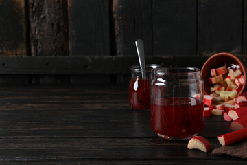 Jars with jam and sliced rhubarb on dark wooden background, space for text