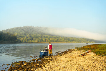 A man, a fisherman in a red vest on the banks of a river, stands next to a motorboat and looks at the morning mist on the river. High quality photo