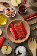 Sliced rhubarb stalk on cutting board, apple, jar of jam, bowls of salt and oil, kitchen utensils on wooden background, top view