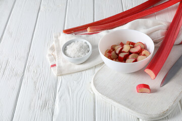 Sliced rhubarb in bowl and on board, knife and bowl of salt on white wooden background, space for text