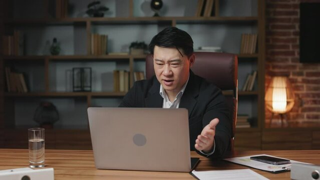 Annoyed office employee working on wireless laptop in office and facing troubles with online program. Asian man of middle age expressing anger and gesturing hands in frustration.