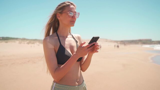 Woman using smartphone standing beach leisure in vacation. Blonde woman in sunglasses, bikini and shorts enjoying sea and texting on cell phone. Social networking on vacation. Using technology outside