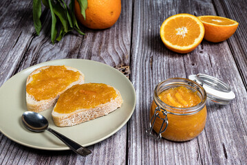Delicious homemade orange marmalade in a glass jar. In the background slices of bread with marmalade in a plate on a white wooden table.