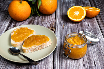 Delicious homemade orange marmalade in a glass jar. In the background slices of bread with marmalade in a plate on a white wooden table.