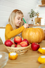 Girl draw on a large orange pumpkin for a Halloween celebration and sit at a wooden table at home. Carved pumpkins into jack-o-lanterns in the kitchen.