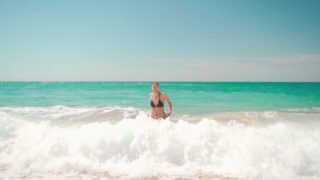 Attractive woman in black bikini comes out water after swimming in ocean. Slim blonde with beautiful body emerges from high waves toward camera. Sensuous woman enjoying her vacation on beach