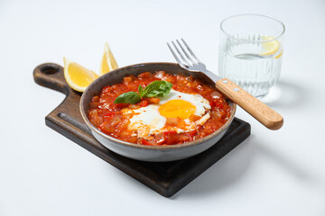 Shakshuka in bowl and fork on board, lemon, glass of water on white background