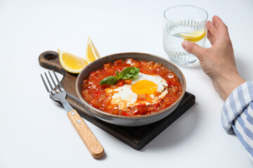 Shakshuka in bowl on board, lemon, glass of water in male hand on white background