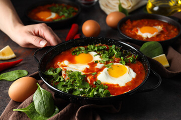 Pans with shakshuka, eggs, basil, bowl of pepper and oil, male hand on dark background