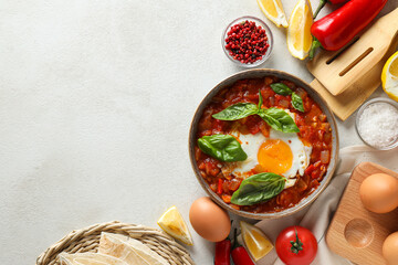 Shakshuka in bowl, spices, tomatoes, eggs and kitchenware on white background, space for text