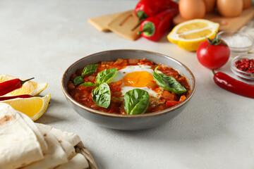 Shakshuka in bowl, spices, tomatoes, eggs and kitchenware on light background
