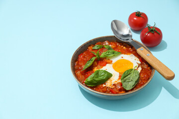 Bowl of shakshuka, spoon and two tomatoes on blue background, space for text
