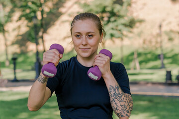 portrait of a young satisfied sports girl doing exercises with dumbbells outdoors during a workout in the park healthy lifestyle concept