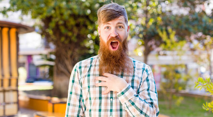 red hair bearded man feeling shocked, astonished and surprised, with hand on chest and open mouth, saying who, me?