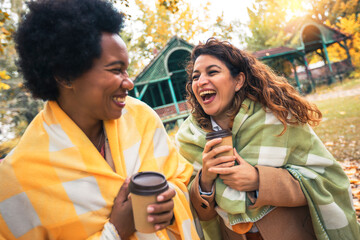 Two smiling young women friends chatting outdoors and drinking coffee while enjoying the walk in the park together