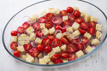 Cherry tomatoes, croutons, red onion, garlic and spices in a glass baking dish on a light gray background, top view. cooking italian panzanella salad