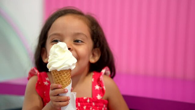 Niña morena comiendo y disfrutando de un delicioso y rico helado para el calor en un fondo rosa en la heladería alegre y contento muy feliz saboreando su nieve en cono