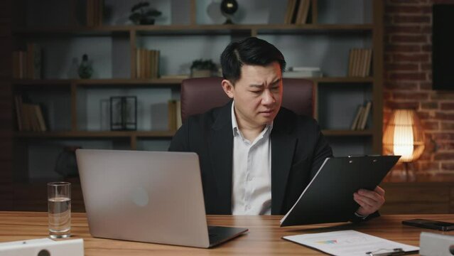 Precise asian supervisor finding out mismatches between paper documents and electronic version. Busy professional spending evening in office working with portable computer.