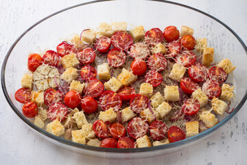 Baked cherry tomatoes, croutons, red onion, garlic, spices and cheese in a glass baking dish on a light gray background, top view. cooking italian panzanella salad