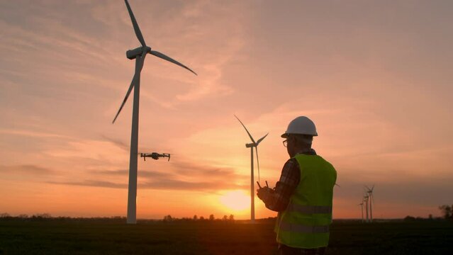 Wind Turbine Specialist, Controls a Flying Drone with a Remote Control, an Engineer in a Helmet and Uniform Checks the Turbines for Operation, Working at a Wind Farm Under Sunrise. Modern Technologies