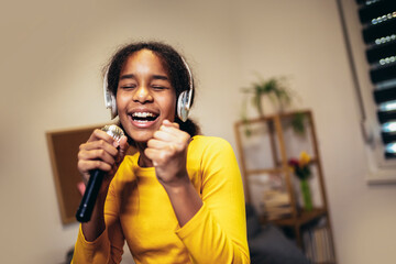 Cute preteen black girl holding a microphone singing karaoke at home, recording songs for a...