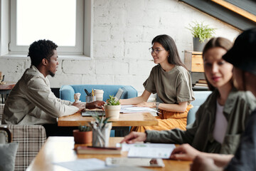 Focus on two young intercultural brokers looking at each other during discussion of financial data while sitting by table in coworking space