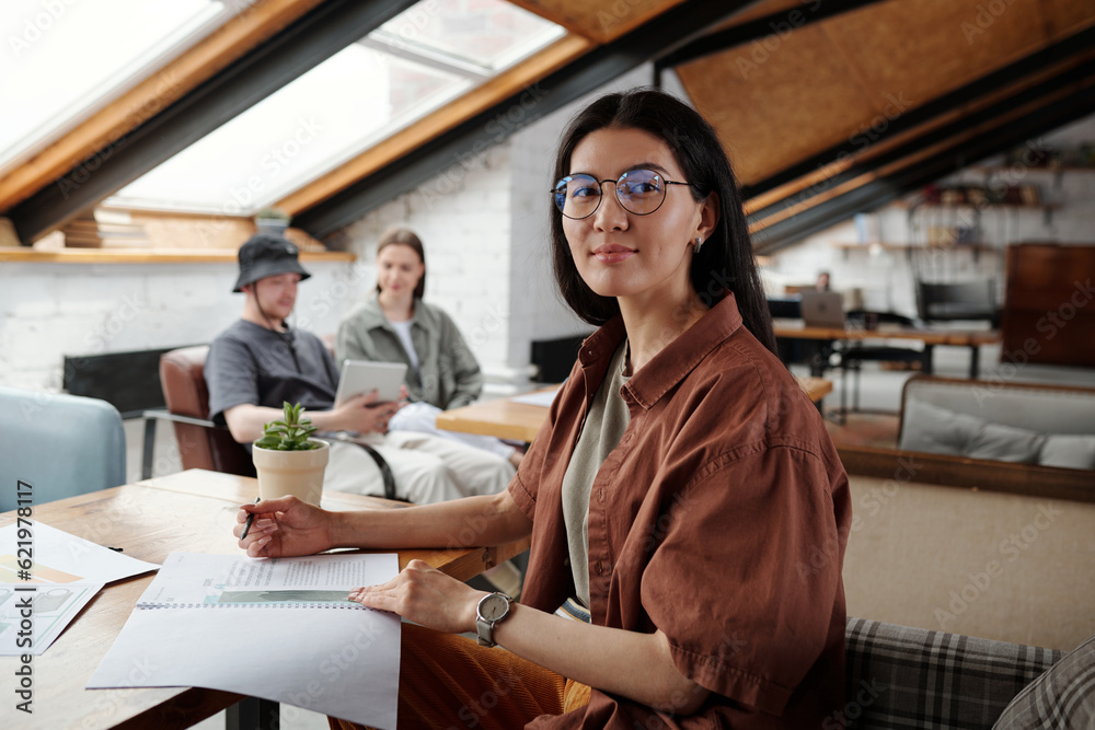 Aufkleber young confident female accountant in eyeglasses sitting by workplace with financial documents and lo
