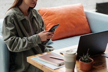 Cropped shot of young businesswoman in grey shirt texting in smartphone while sitting on comfortable couch by workplace in front of laptop