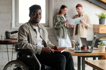 Happy young African American male broker with disability sitting in wheelchair in front of camera...