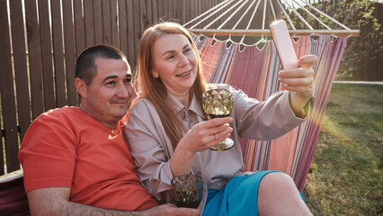 Happy married couple sitting in a hammock drinking wine and taking selfies during a family dinner in the backyard of the house