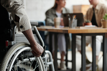 Part of young African American male employee with disability keeping hand on wheel of wheelchair...