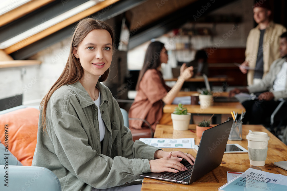 Poster young smiling female analyst or economist looking at camera while sitting by workplace in front of l