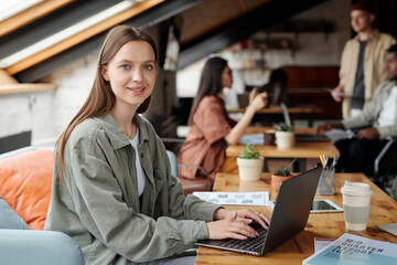 Young smiling female analyst or economist looking at camera while sitting by workplace in front of...