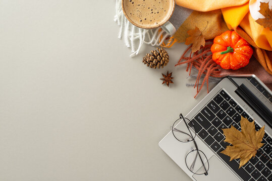 Embodying the concept of working from home, an above view image showcases a laptop amidst a cozy blanket, pumpkin, pinecone, glasses, hot chocolate on isolated grey background with copyspace