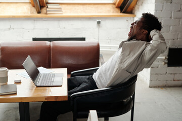 Side view of young yawning businessman with hands behind his head sitting in armchair by workplace...