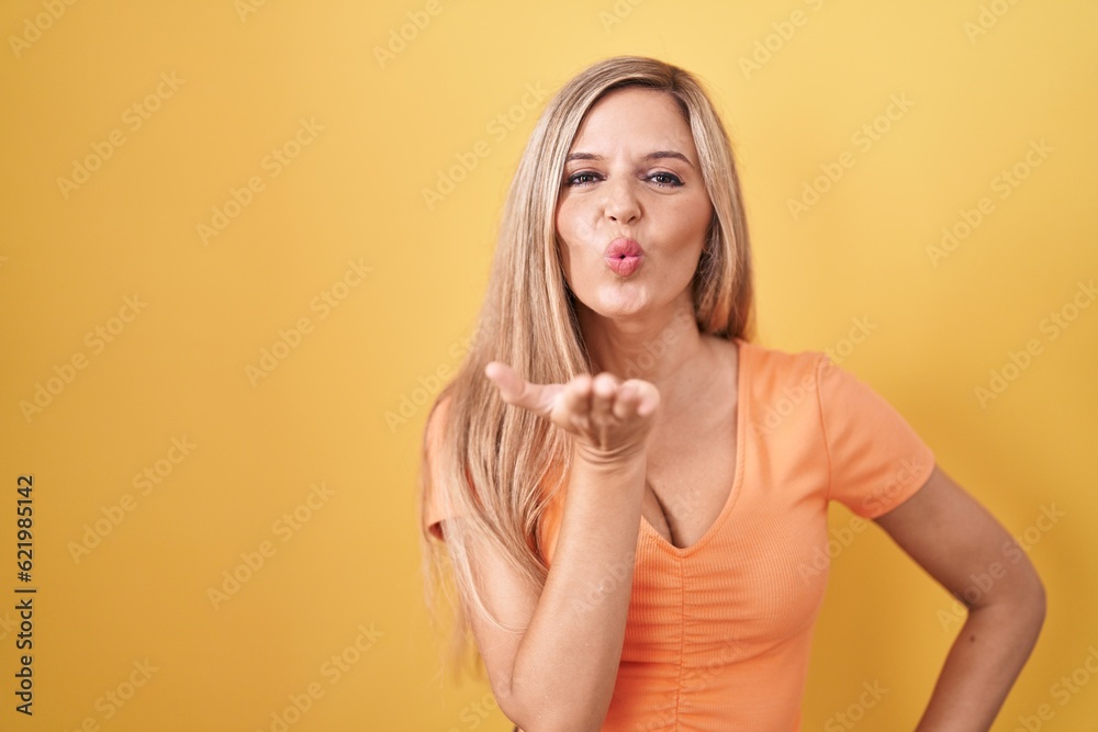 Poster young woman standing over yellow background looking at the camera blowing a kiss with hand on air be