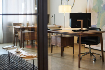 Part of coworking area with wooden desks, computer monitor, lamp, armchair, long small table for having lunch and wooden cabinet