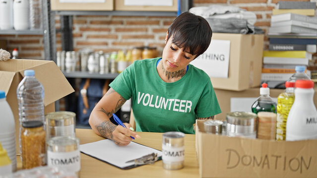 Young beautiful hispanic woman volunteer sitting on table writing on clipboard at charity center