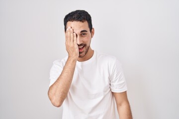 Handsome hispanic man standing over white background covering one eye with hand, confident smile on...