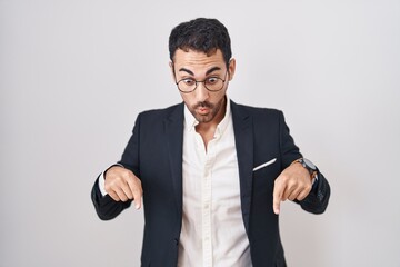 Handsome business hispanic man standing over white background pointing down with fingers showing advertisement, surprised face and open mouth